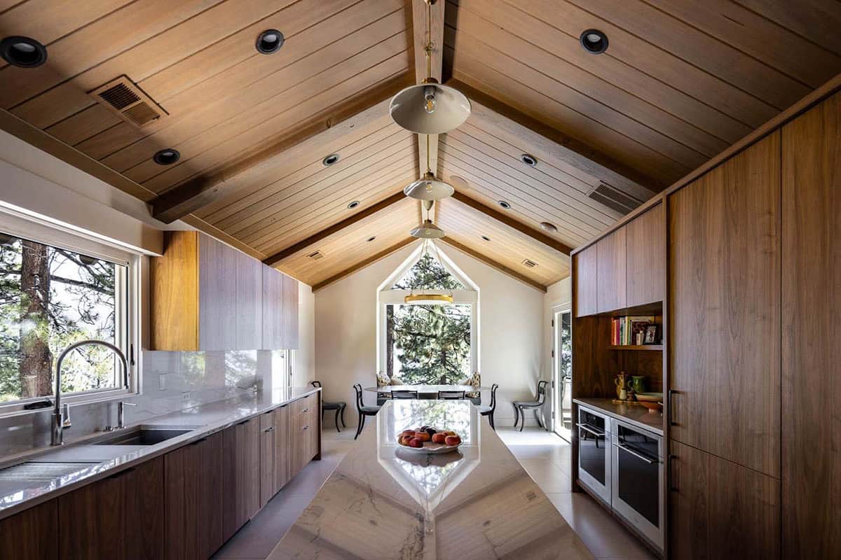Kitchen looking toward breakfast nook with arched gable window framing pine trees and lake, walnut cabinetry and marble island