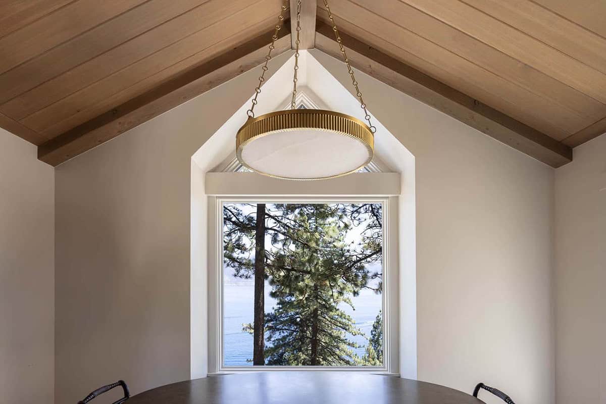 Dining nook detail with brass ring pendant, arched gable window framing pine trees and glimpse of Lake Tahoe beyond
