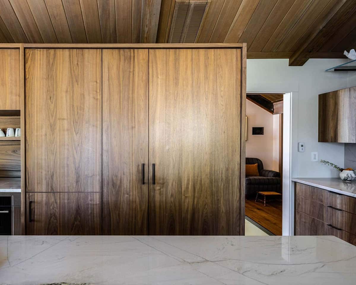 Floor-to-ceiling walnut pantry cabinet with black hardware, marble island in foreground, opening to living room beyond