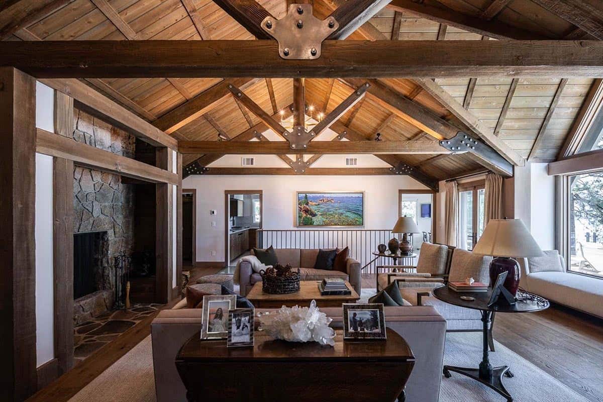 Lodge-style great room with soaring timber truss ceiling, iron hardware, stone fireplace, and lake views through large windows