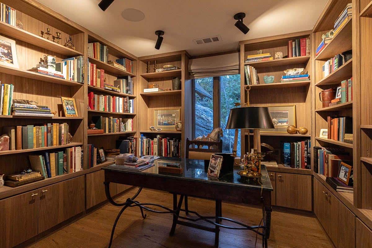 Cozy wood-paneled home library with floor-to-ceiling bookshelves, antique iron desk, brass lamp, and forest window view