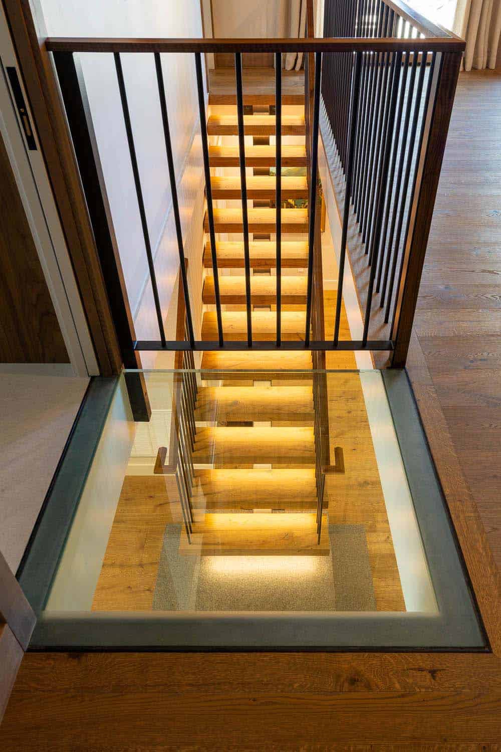 Bird's-eye view looking down through glass floor panel at illuminated floating staircase below