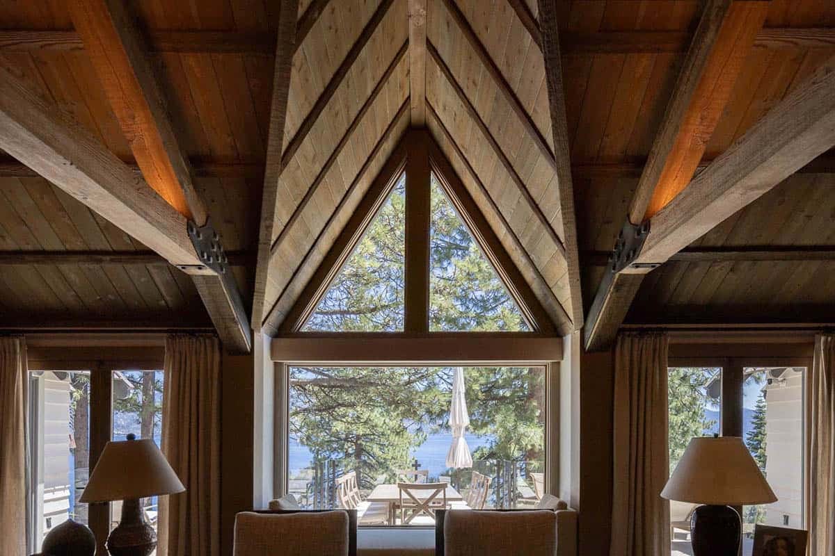 Dramatic timber A-frame gable window framing pine trees and Lake Tahoe, flanked by rustic beam ceiling and table lamps