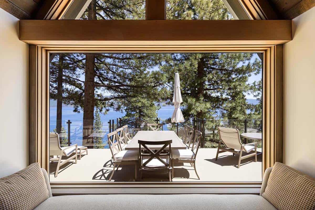 View from living room window seat through triangular gable window to lakeside terrace with dining set and Lake Tahoe beyond