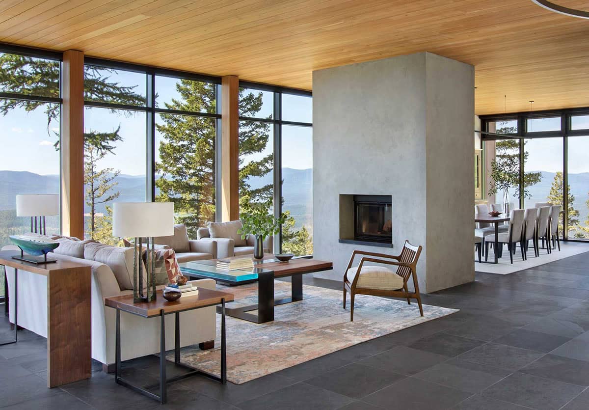 Open-plan living and dining room with concrete fireplace, wood ceiling, and panoramic forest views