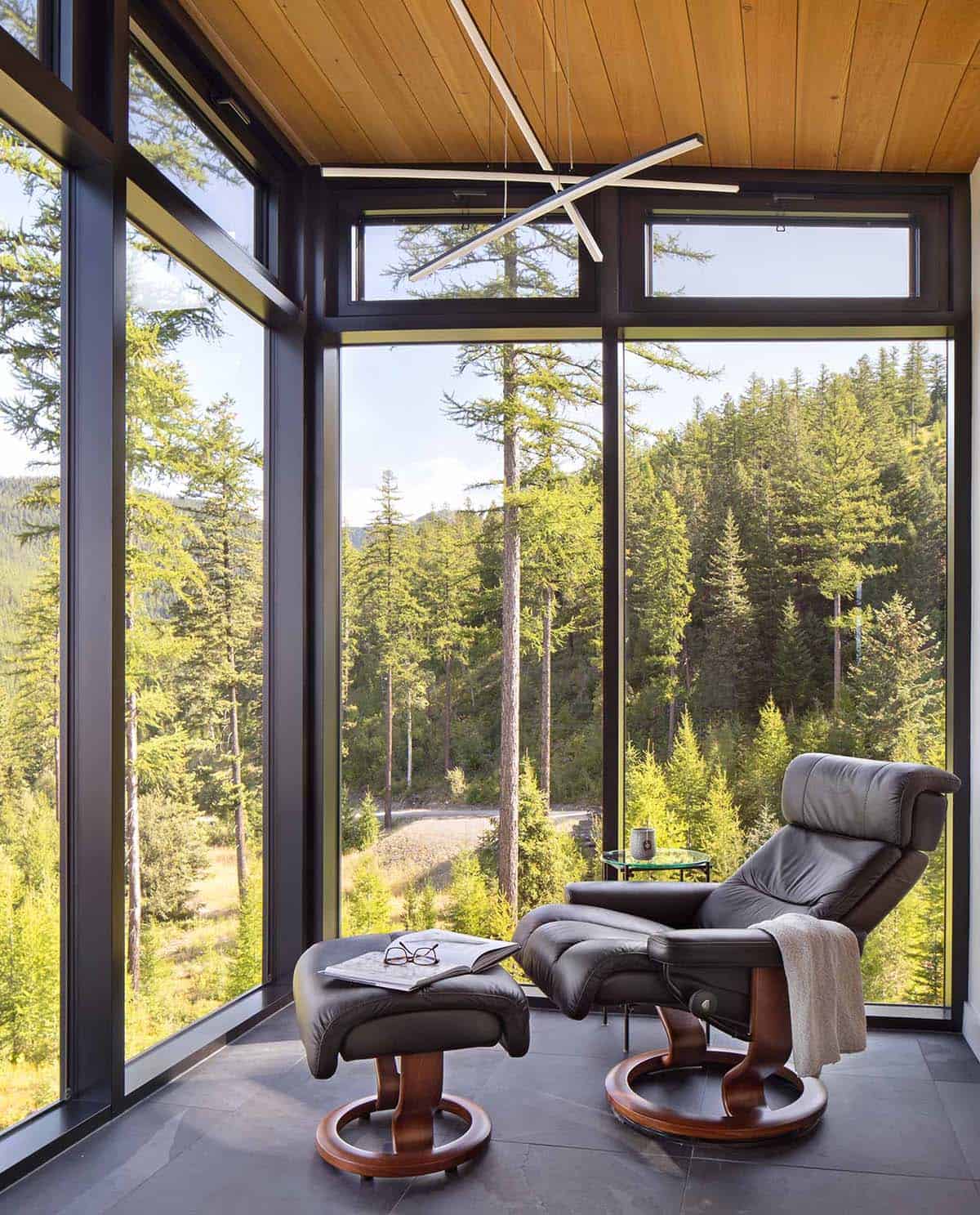 Leather lounge chair and ottoman in glass corner reading nook surrounded by forest treetops