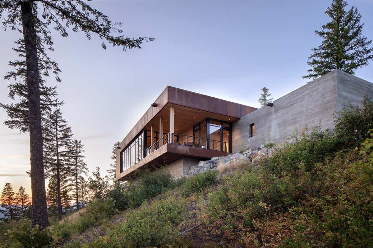 Corten steel and concrete home cantilevered over rocky cliff at twilight among pine trees
