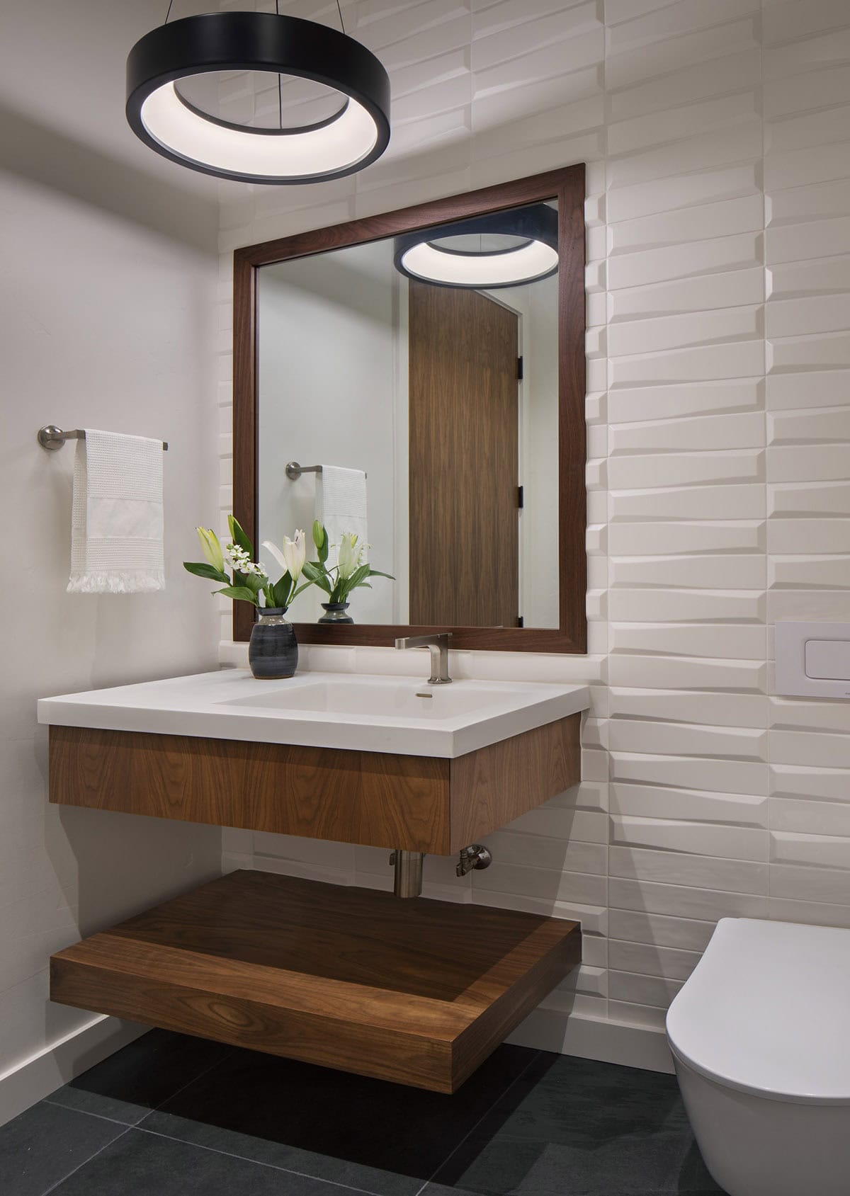 Floating walnut vanity with white stone countertop, framed mirror, and textured white tile wall