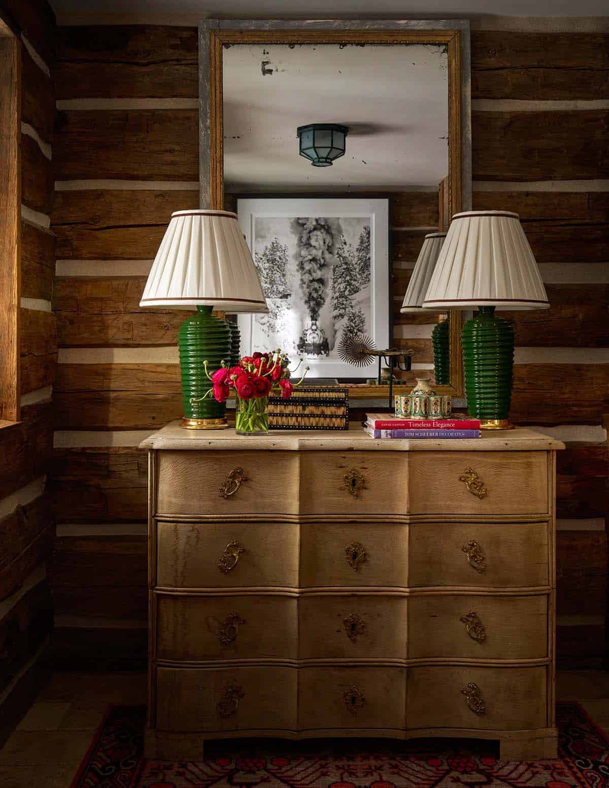 Antique chest with pair of green ceramic lamps, red ranunculus, and antiqued mirror against log wall