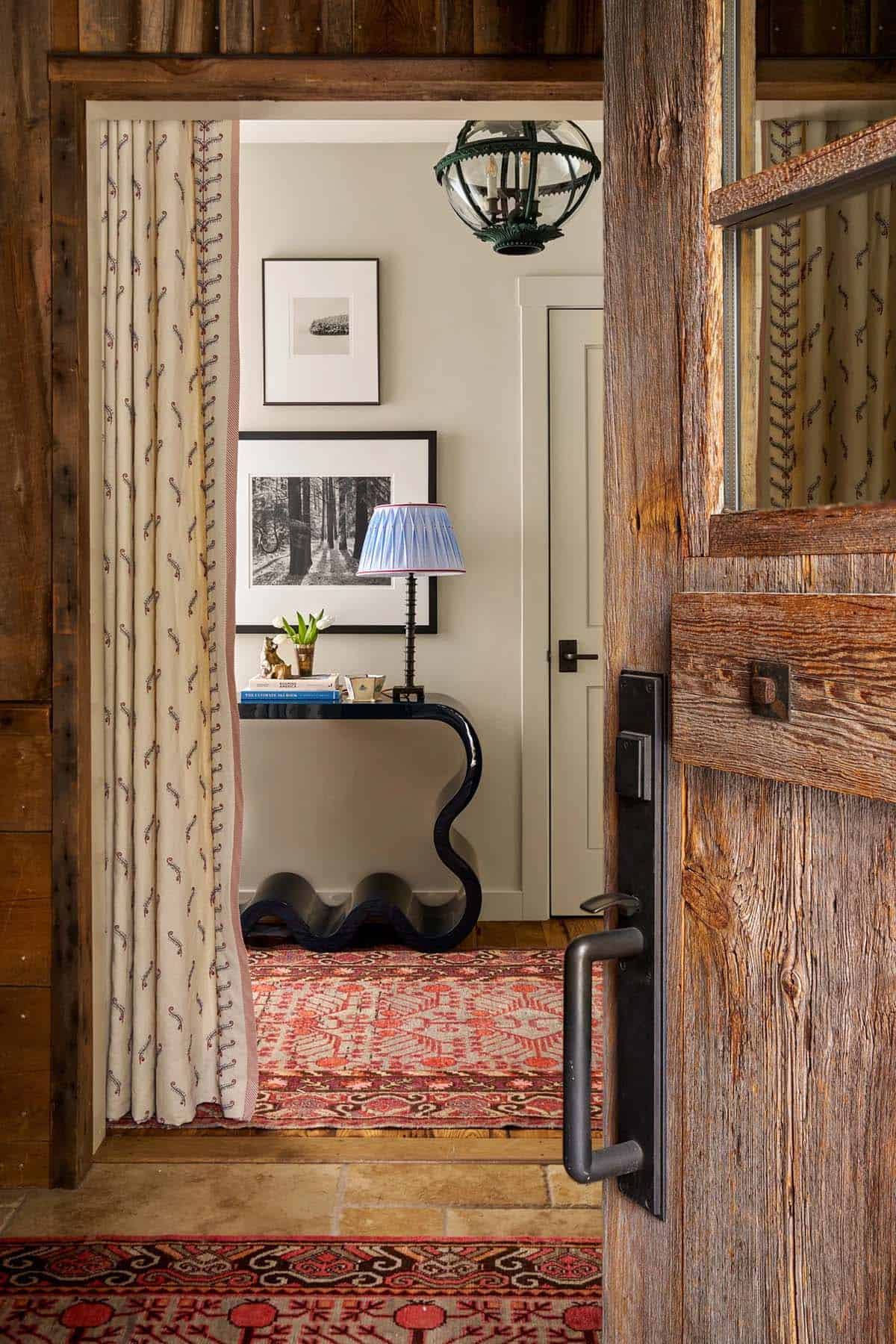 View through rustic timber door into hallway with wavy black console table, globe pendant, and printed curtains