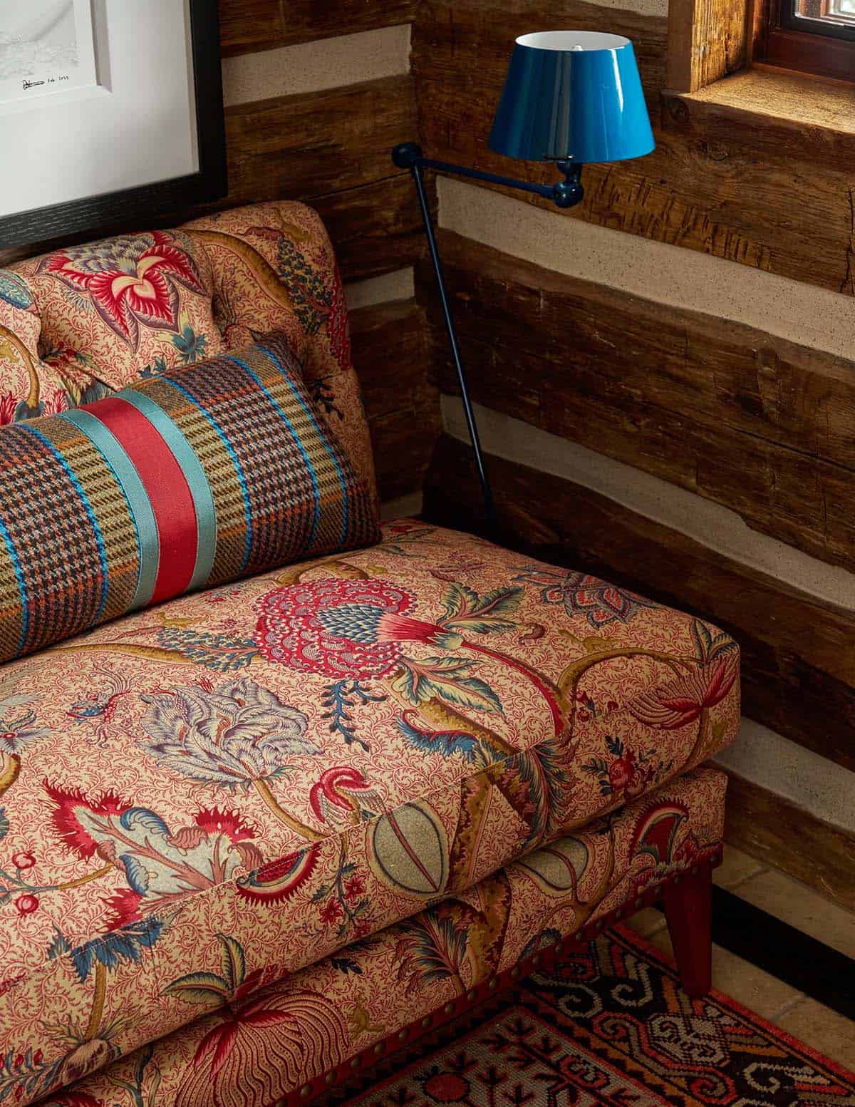 Close-up of richly patterned floral daybed with plaid bolster pillow and blue swing-arm lamp against log wall