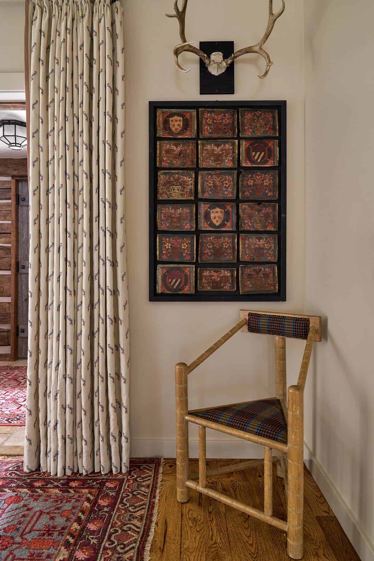 Hallway corner with antler mount, framed heraldic tile collection, houndstooth chair, and printed curtains