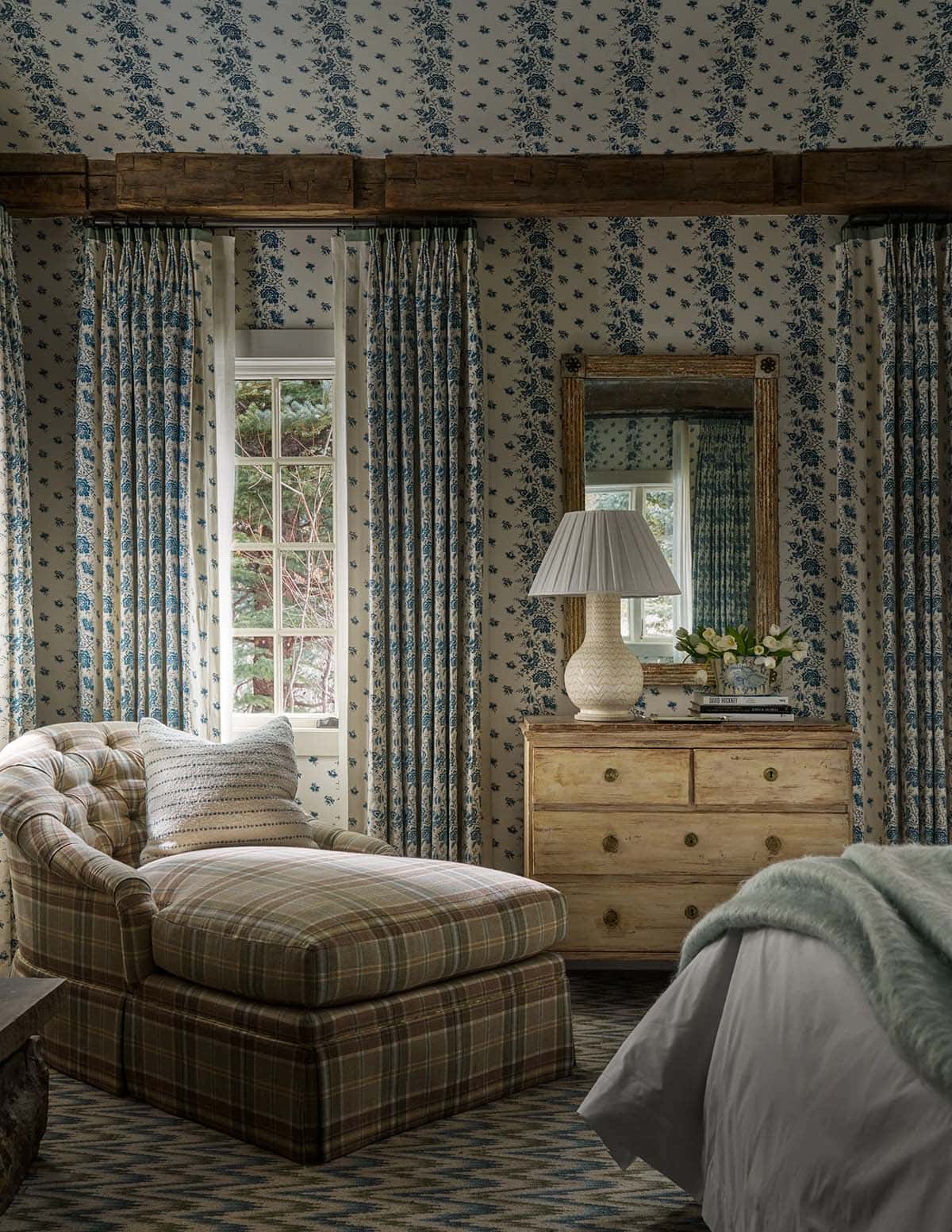 Guest bedroom wrapped in blue floral wallpaper with plaid chaise, matching curtains, and antique pine dresser