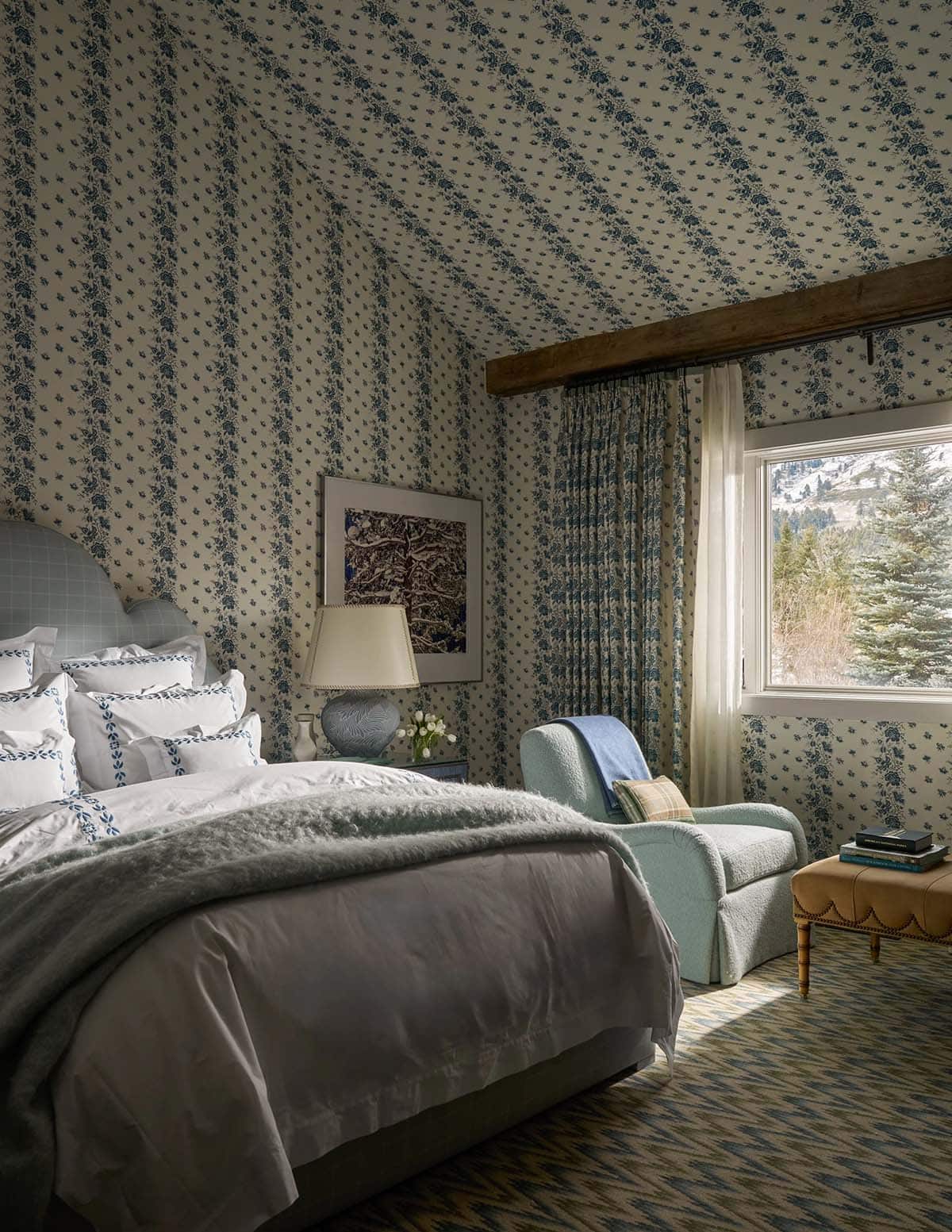 Blue floral wallpapered bedroom with upholstered headboard, plaid chaise, and snow-capped mountain view