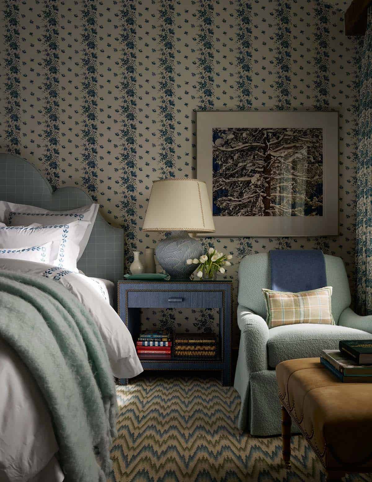 Bedroom detail with blue floral wallpaper, navy nightstand, plaid armchair, and winter tree artwork