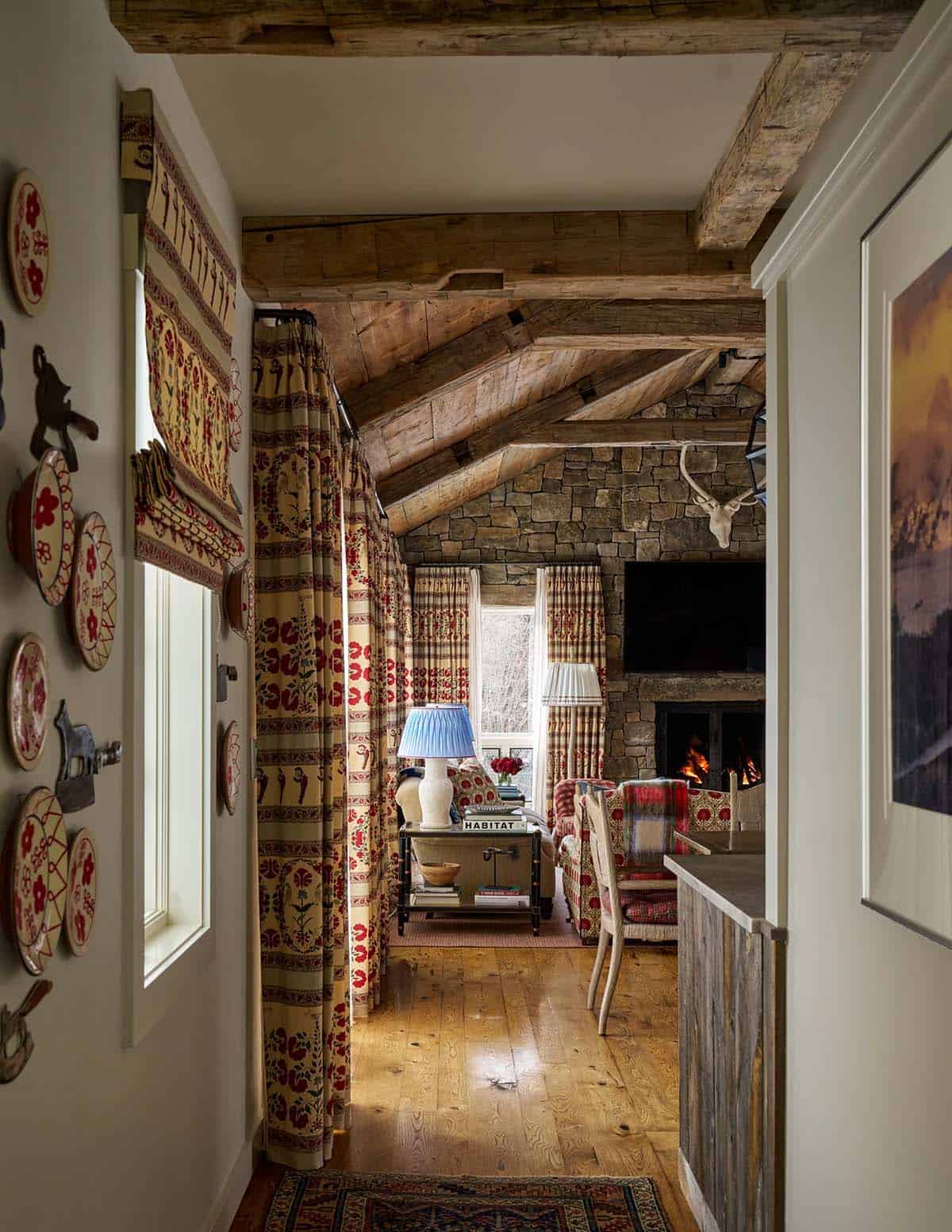 Hallway view into family room with vaulted timber ceiling, stone fireplace, and colorful patterned curtains