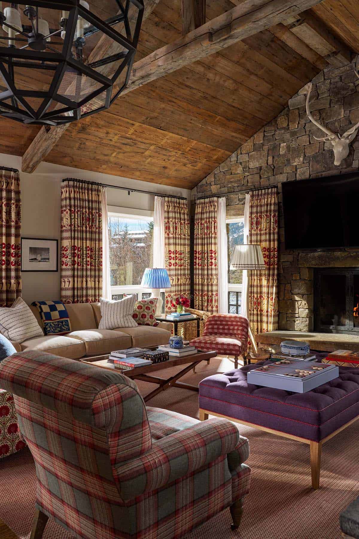 Wide view of family room with vaulted timber ceiling, stone fireplace, plaid armchairs, and geometric chandelier