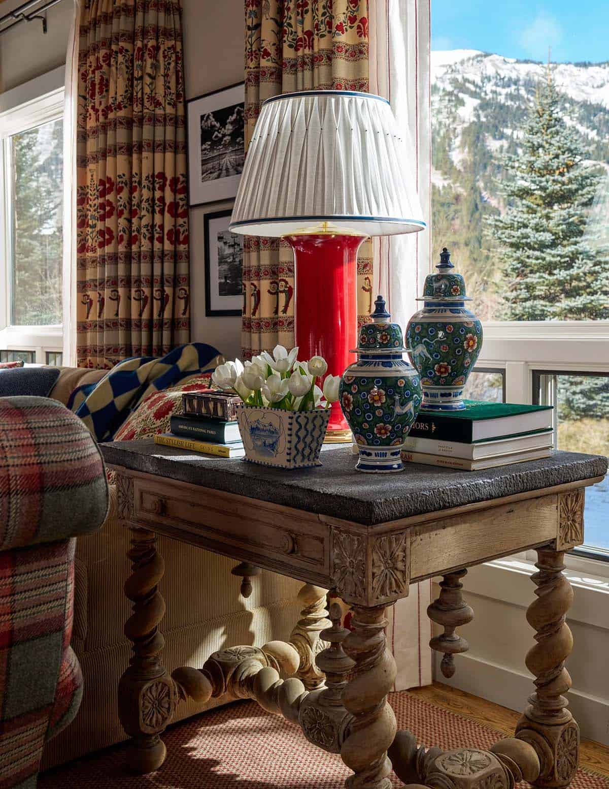 Carved antique side table with red lamp, ginger jars, and white tulips against snow-covered mountain view