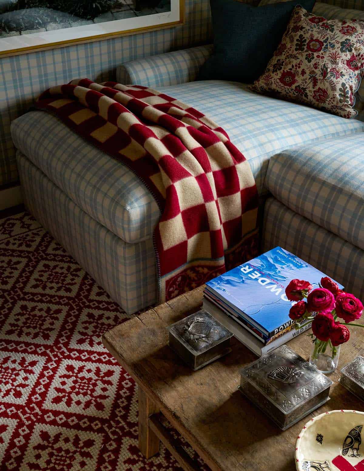 Close-up of plaid sofa with red checkered throw, rustic coffee table, ski books, and red Nordic rug