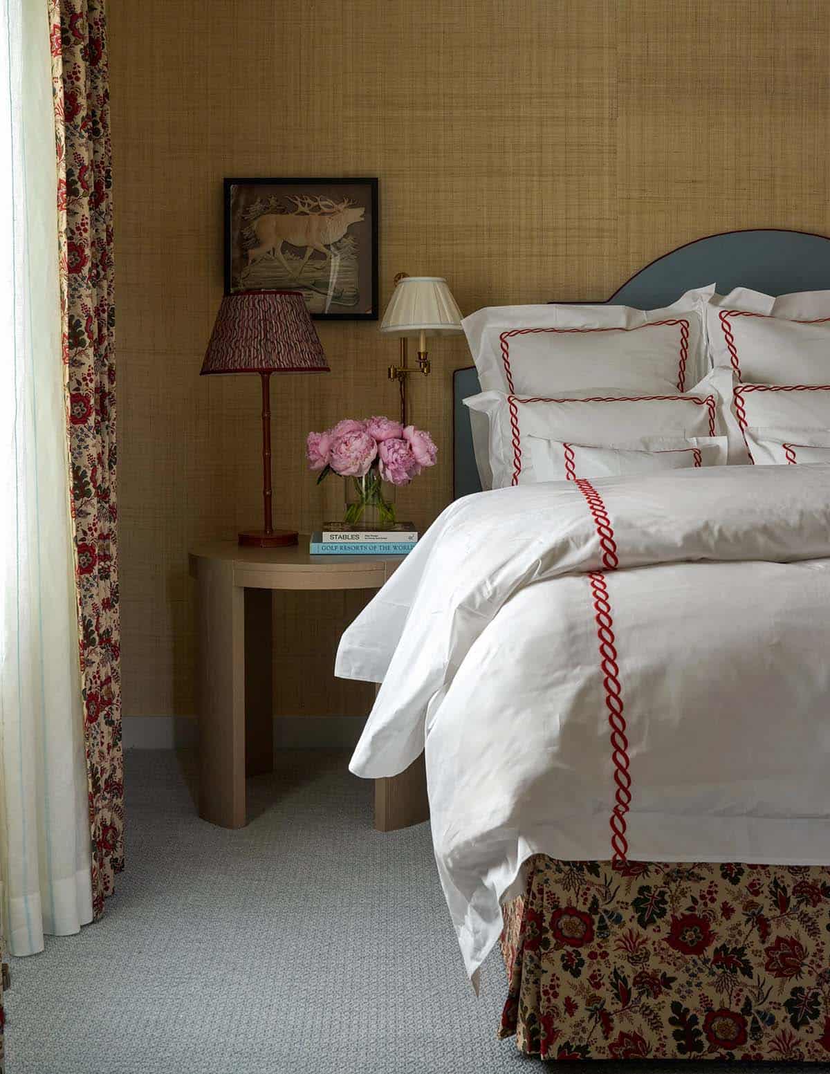 Guest bedroom detail with blue upholstered headboard, red-trimmed white linens, grasscloth walls, and pink peonies