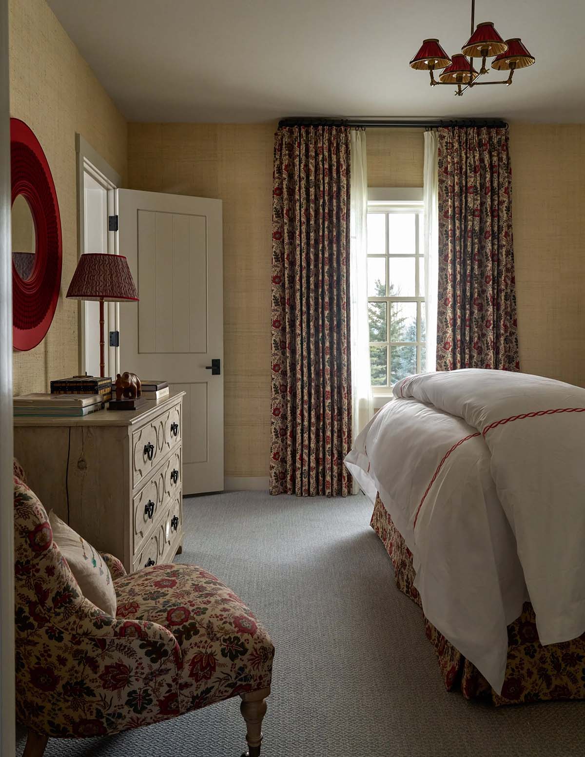 Guest bedroom with grasscloth walls, floral curtains, white chest, red lamp, and red-trimmed bedding