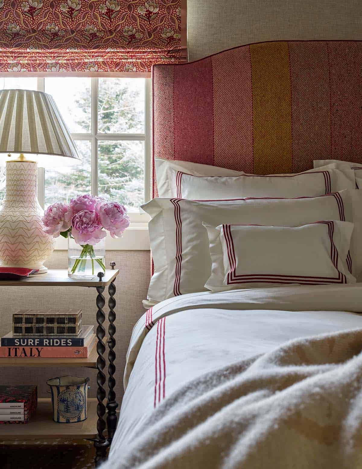 Close-up of striped upholstered headboard with red-trimmed white linens, pink peonies, and barley twist nightstand