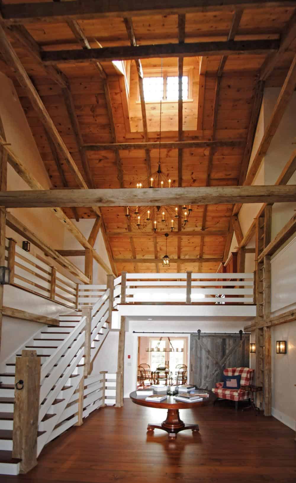 Barn house foyer with exposed timber frame ceiling, white staircase, mezzanine loft, and sliding barn door