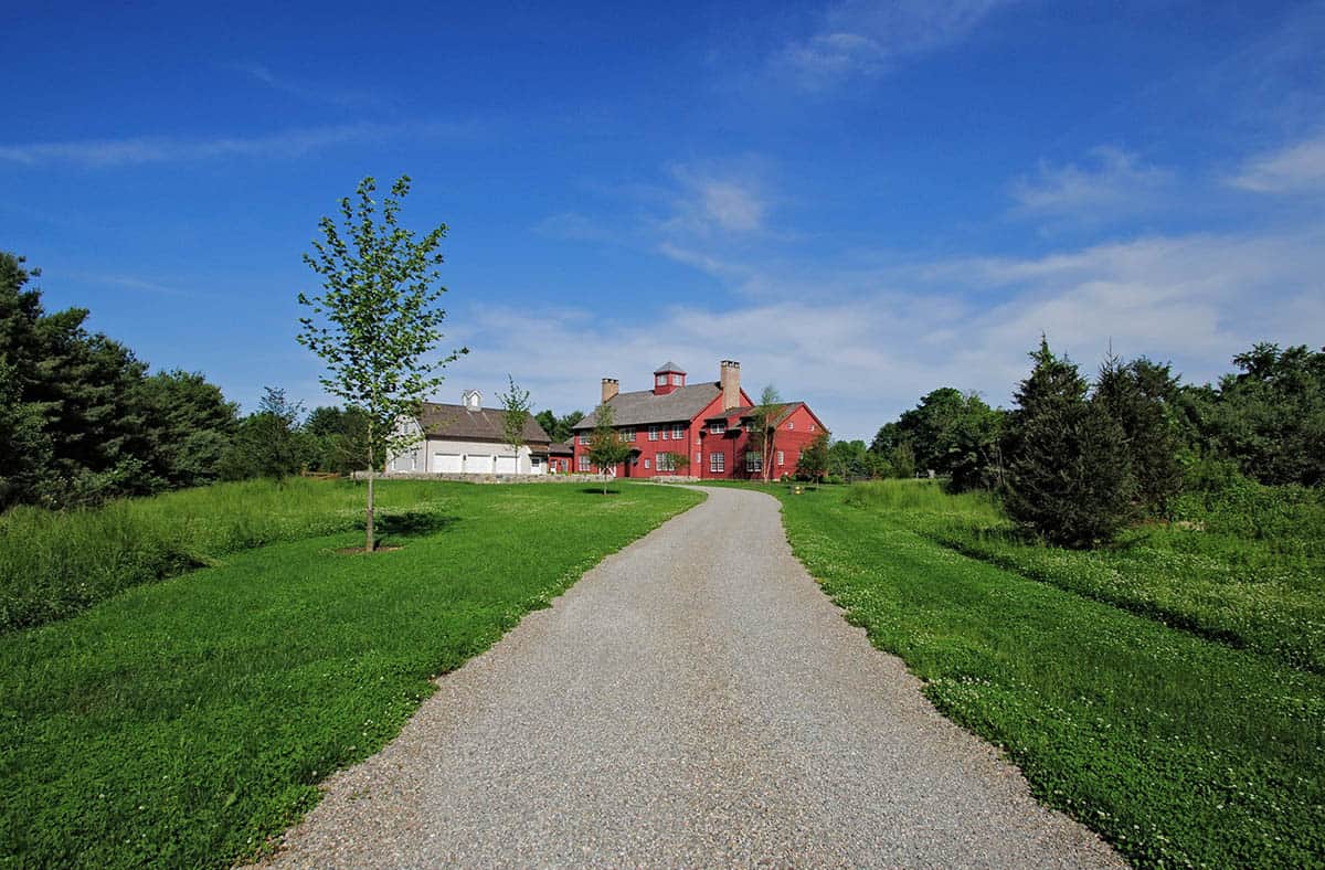 Long gravel driveway leading to red New England barn house surrounded by trees and green meadow