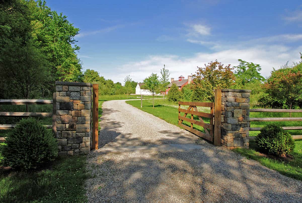 Stone pillar and wood farm gate entrance with gravel driveway leading to red barn house