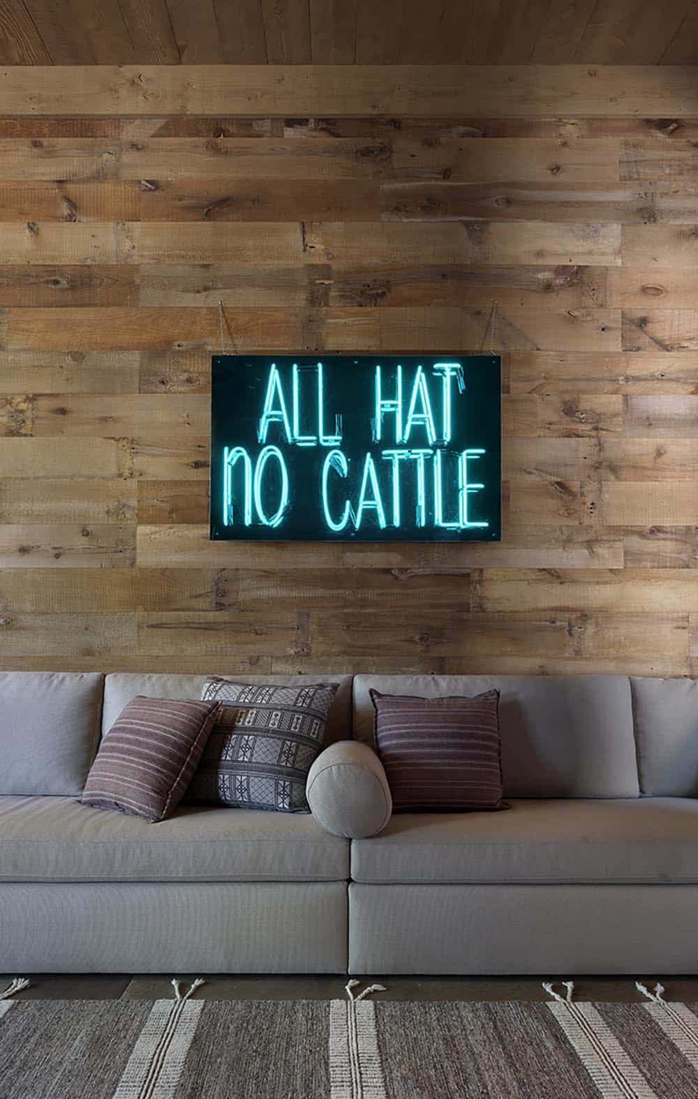 Neon sign reading All Hat No Cattle on reclaimed wood wall above gray sofa