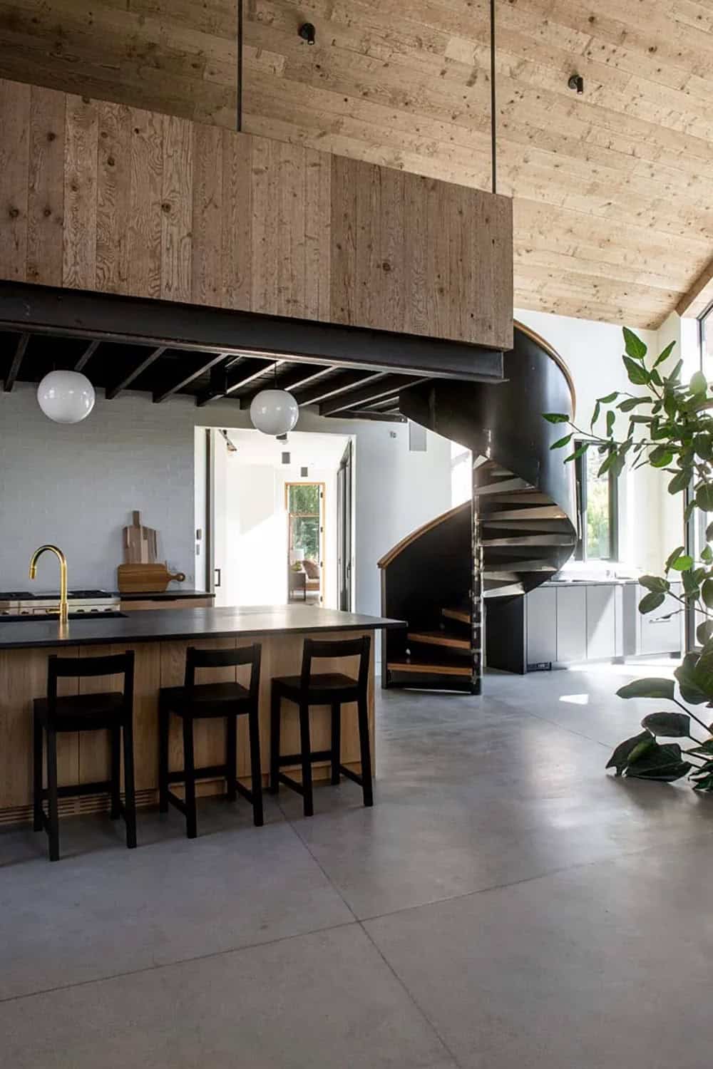 Kitchen island with wood base and dark countertop alongside dramatic spiral steel staircase and indoor fig tree