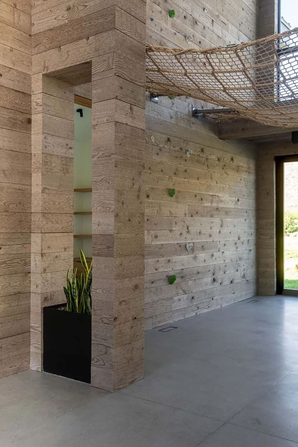 Indoor climbing wall with rope holds and overhead cargo net loft in reclaimed wood-lined recreation room