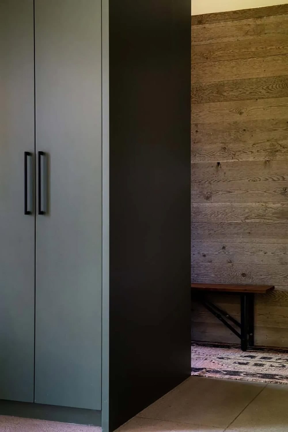 Sage green built-in storage with black steel door opening to reclaimed wood mudroom bench and vintage rug