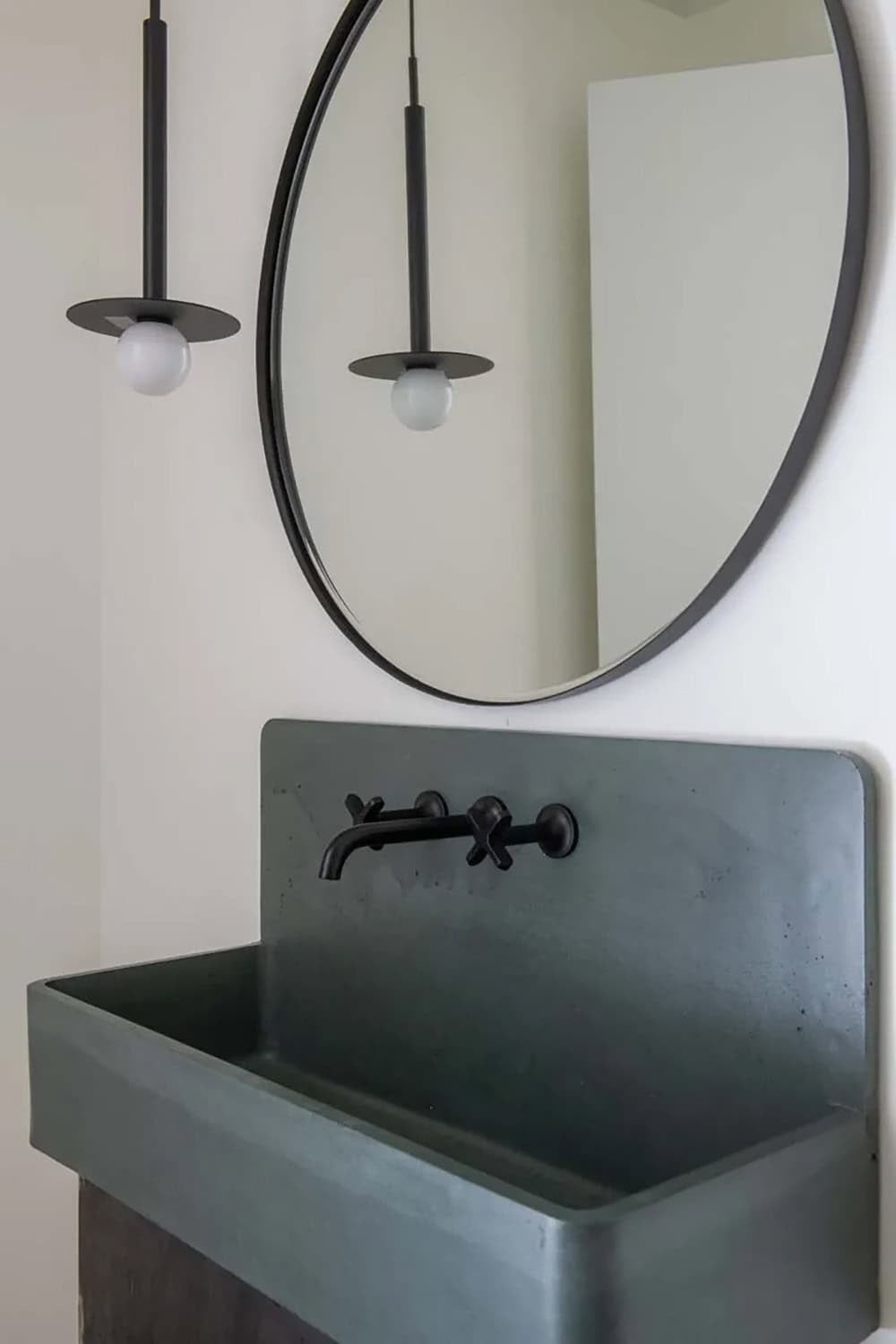Close-up of slate-toned concrete trough sink with matte black wall-mount faucet and round black-framed mirror