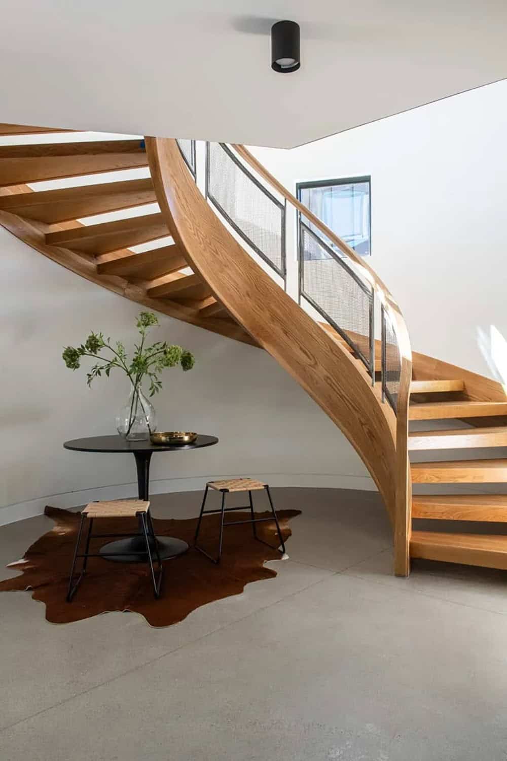 Curved oak staircase with mesh railing, cowhide rug, and tulip table beneath in stone silo interior