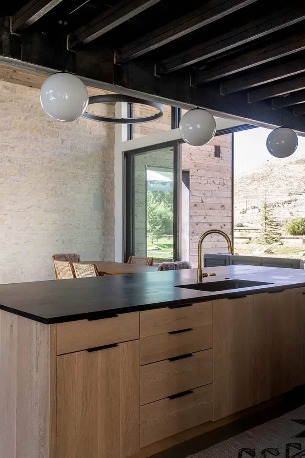 Oak kitchen island with dark stone countertop, brass faucet, globe pendants, and open sliding door to patio