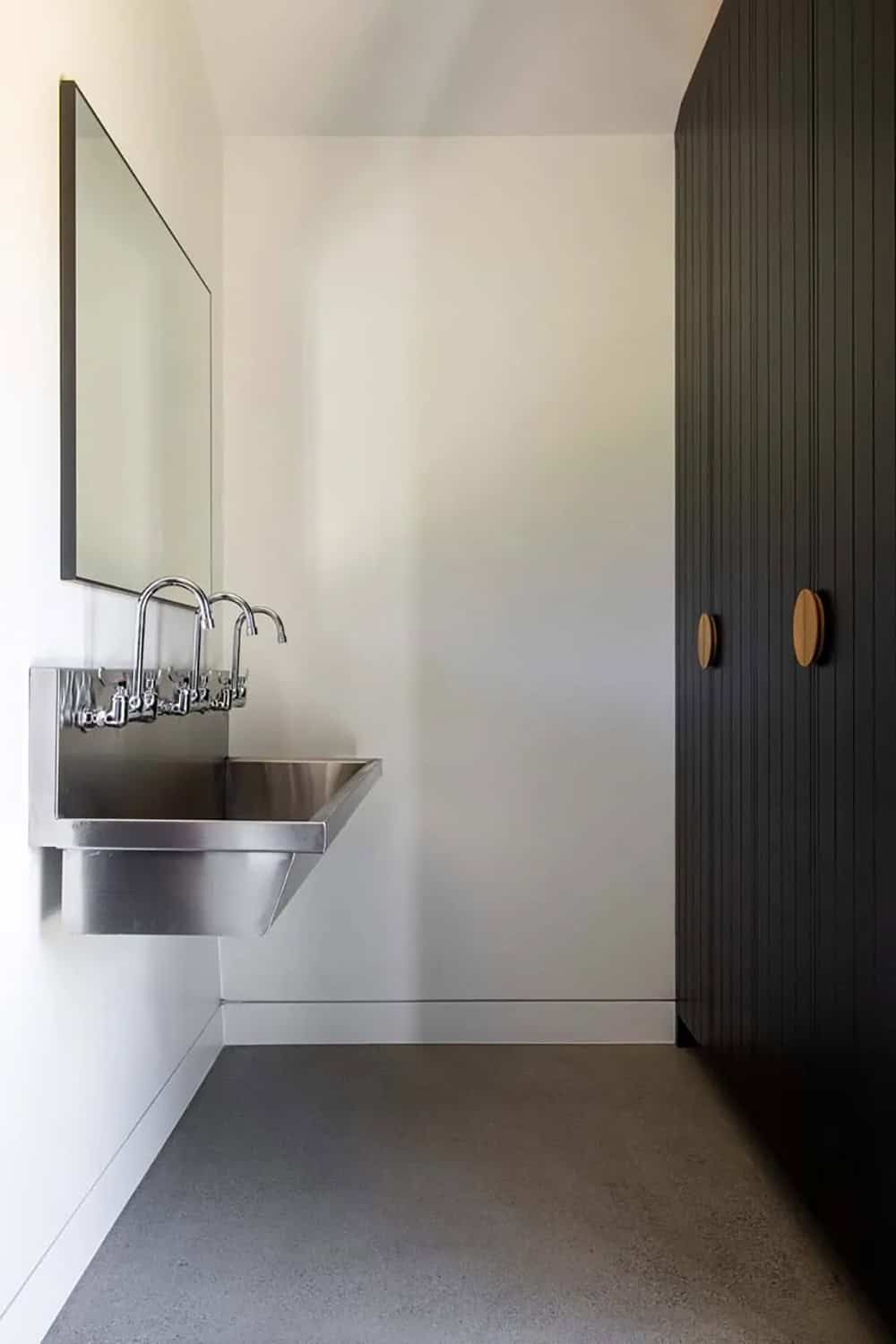 Industrial stainless steel trough sink with dual faucets beside black vertical-board cabinetry with wood pulls