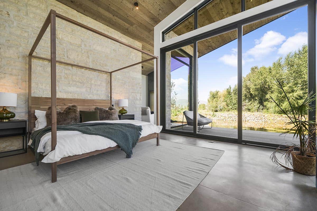 Four-poster walnut bed against limestone feature wall with floor-to-ceiling glazing opening to pond patio, Park City