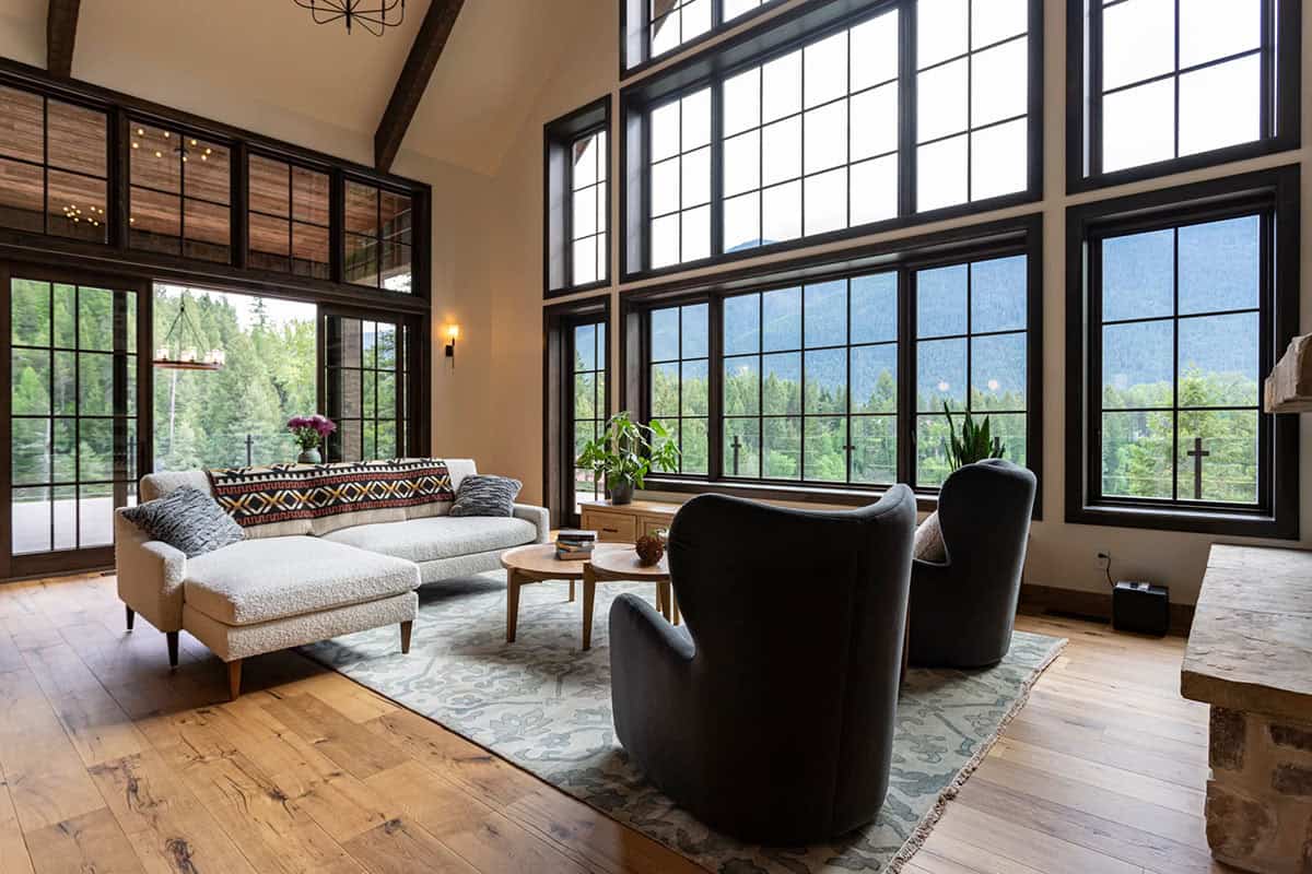 Soaring great room with floor-to-ceiling black-framed windows framing mountain views, white oak floors, and a mix of boucle and dark accent chairs