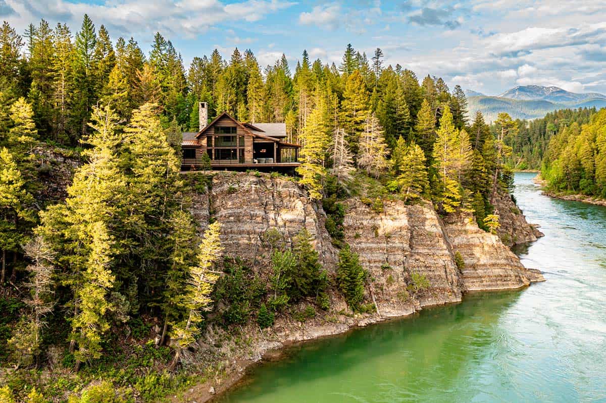 Aerial view of River Dance log cabin nestled in pine forest atop a layered rock cliff above the turquoise Flathead River.