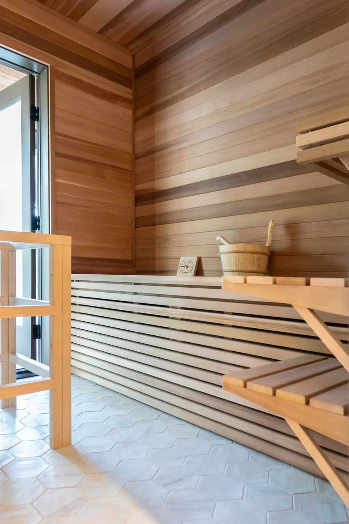 Custom cedar sauna with tiered wood benches, a wooden bucket, and hexagonal white tile floors