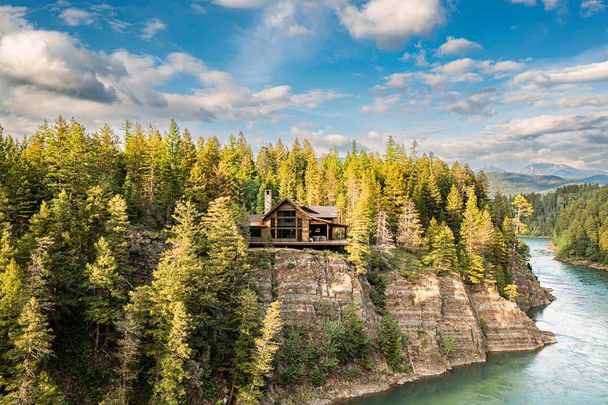 Wide aerial view of River Dance cabin on a forested cliff overlooking the winding turquoise Flathead River with mountains in the distance
