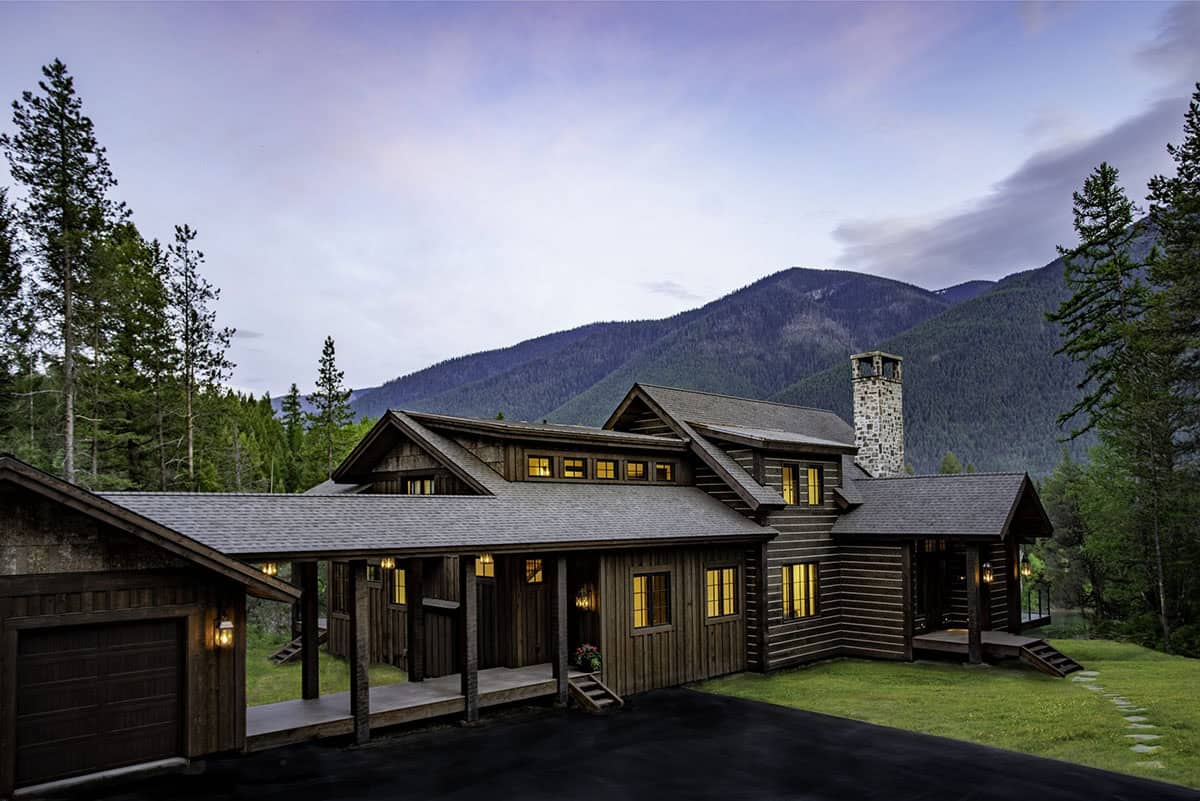 Rear exterior of River Dance at dusk with covered breezeway, stone chimney, log cabin siding, and mountain backdrop