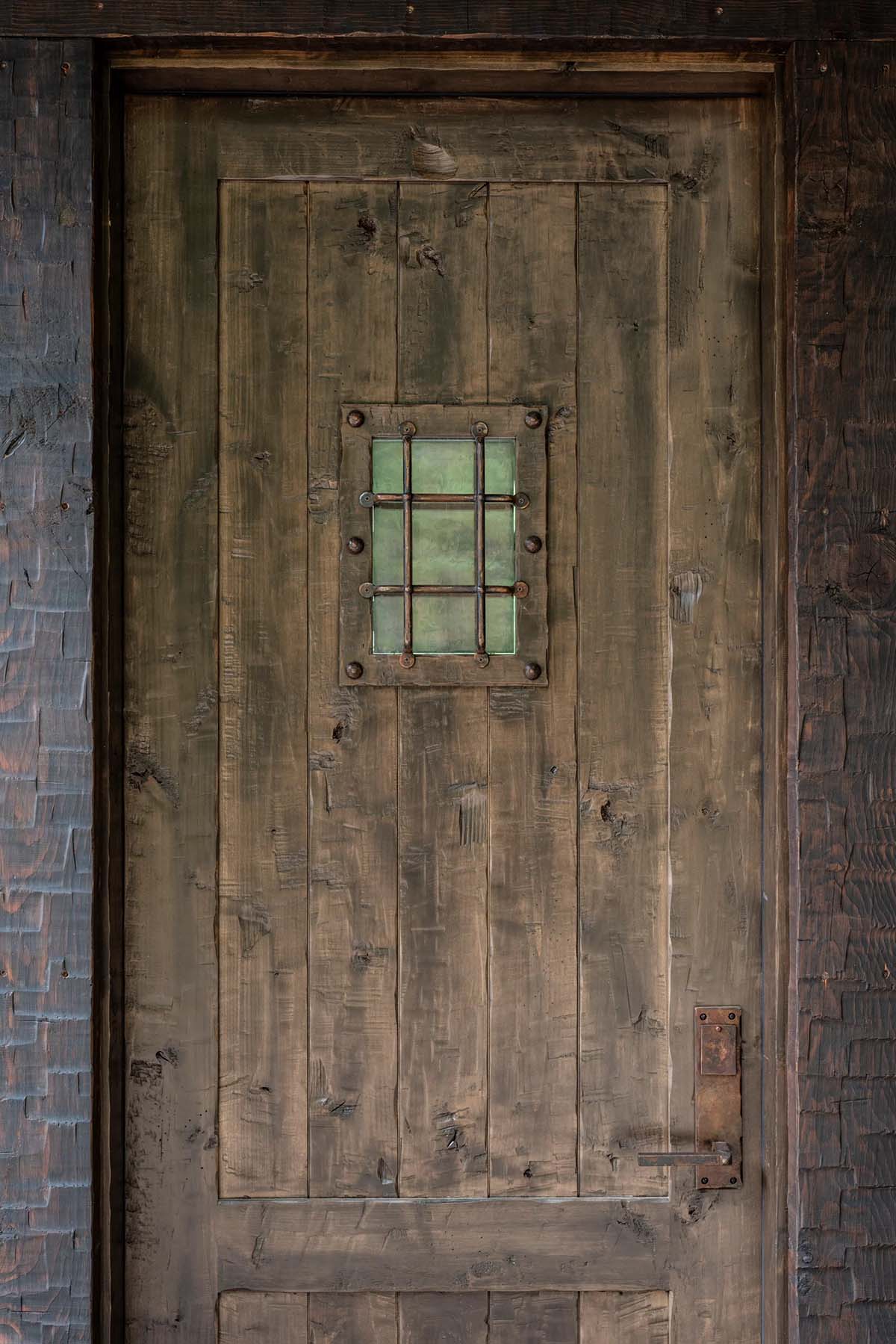 Rustic hand-hewn entry door with a small iron-barred green glass window set into salvaged poplar bark siding