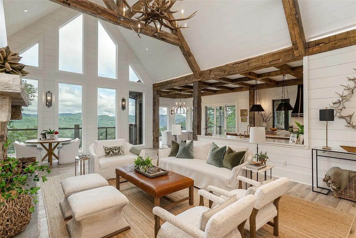 Rustic mountain home great room with white slipcovered sofas, antler chandelier, exposed wood beams, and floor-to-ceiling mountain views