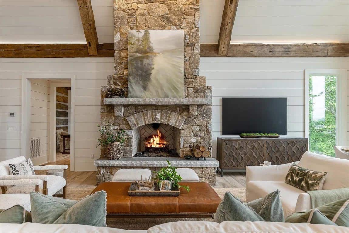 Stone fireplace with arched opening and active fire, flanked by TV cabinet and white shiplap walls with reclaimed beam ceiling