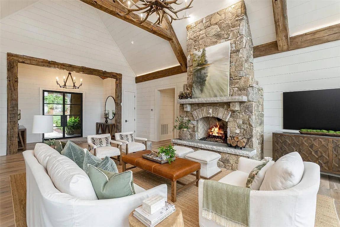Great room with stone fireplace, antler chandelier, vaulted shiplap ceiling, reclaimed wood beam doorway, and white slipcovered seating