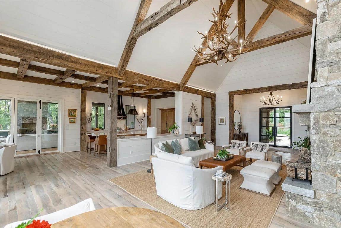 Open-concept living and kitchen area with reclaimed wood beams, antler chandelier, white shiplap walls, and view to outdoor fireplace