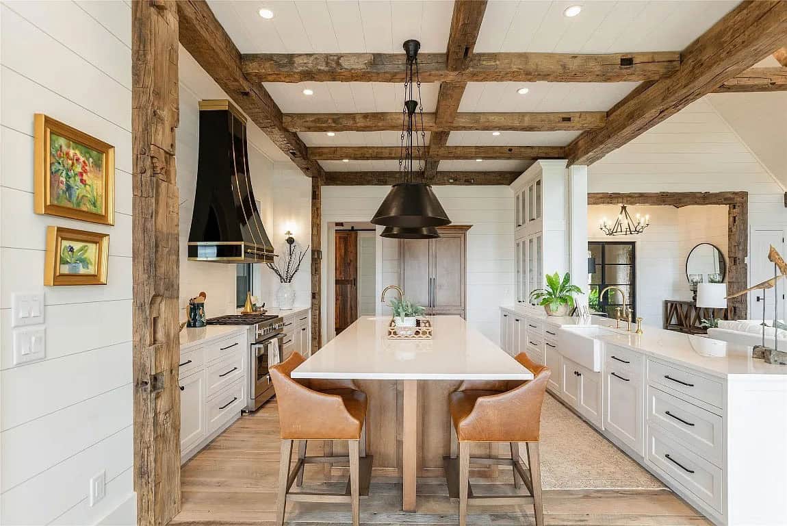 Kitchen with reclaimed beam ceiling, white cabinetry, wood island with leather stools, black and brass range hood, and farmhouse sink