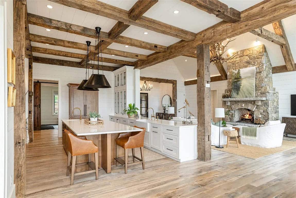 Open kitchen and living area with reclaimed timber posts, white island, stone fireplace, and antler chandelier in background